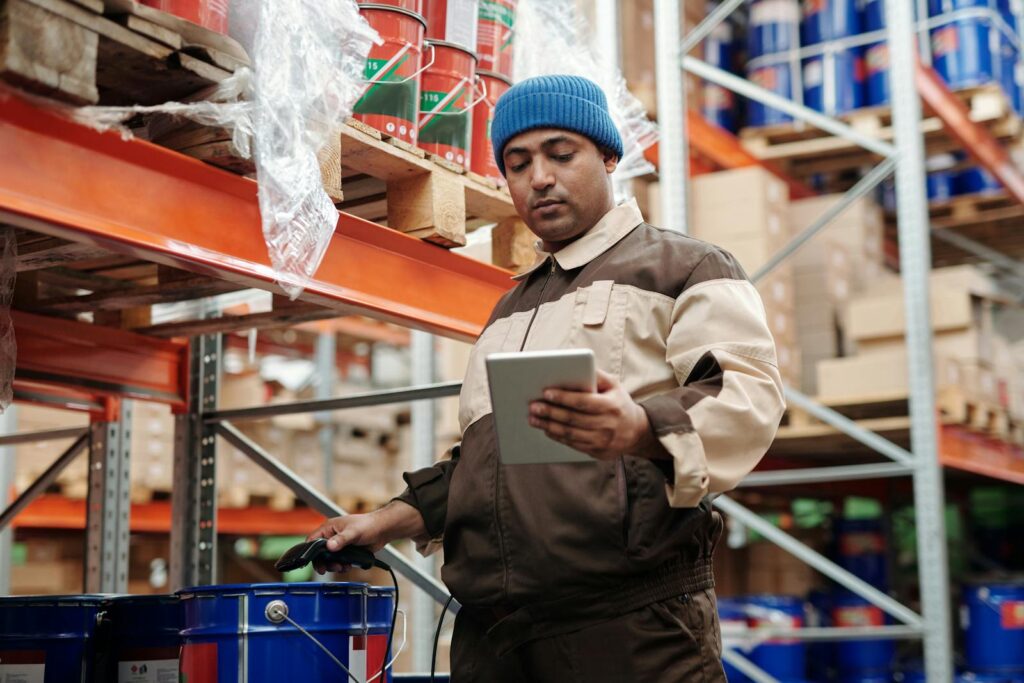 Man in blue beanie uses tablet in a warehouse for inventory management.