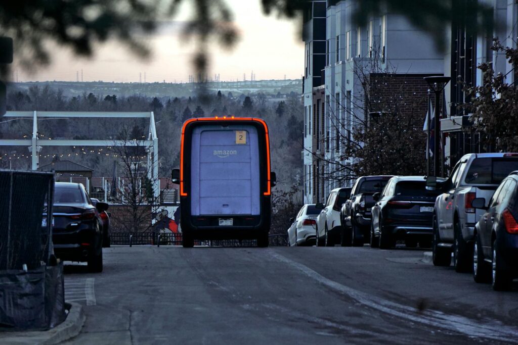 Amazon delivery van driving through an urban street at twilight with cars parked along the side.