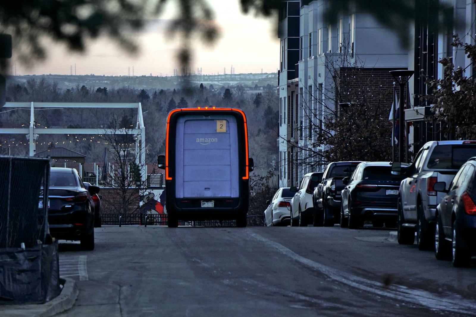 Amazon delivery van driving through an urban street at twilight with cars parked along the side.