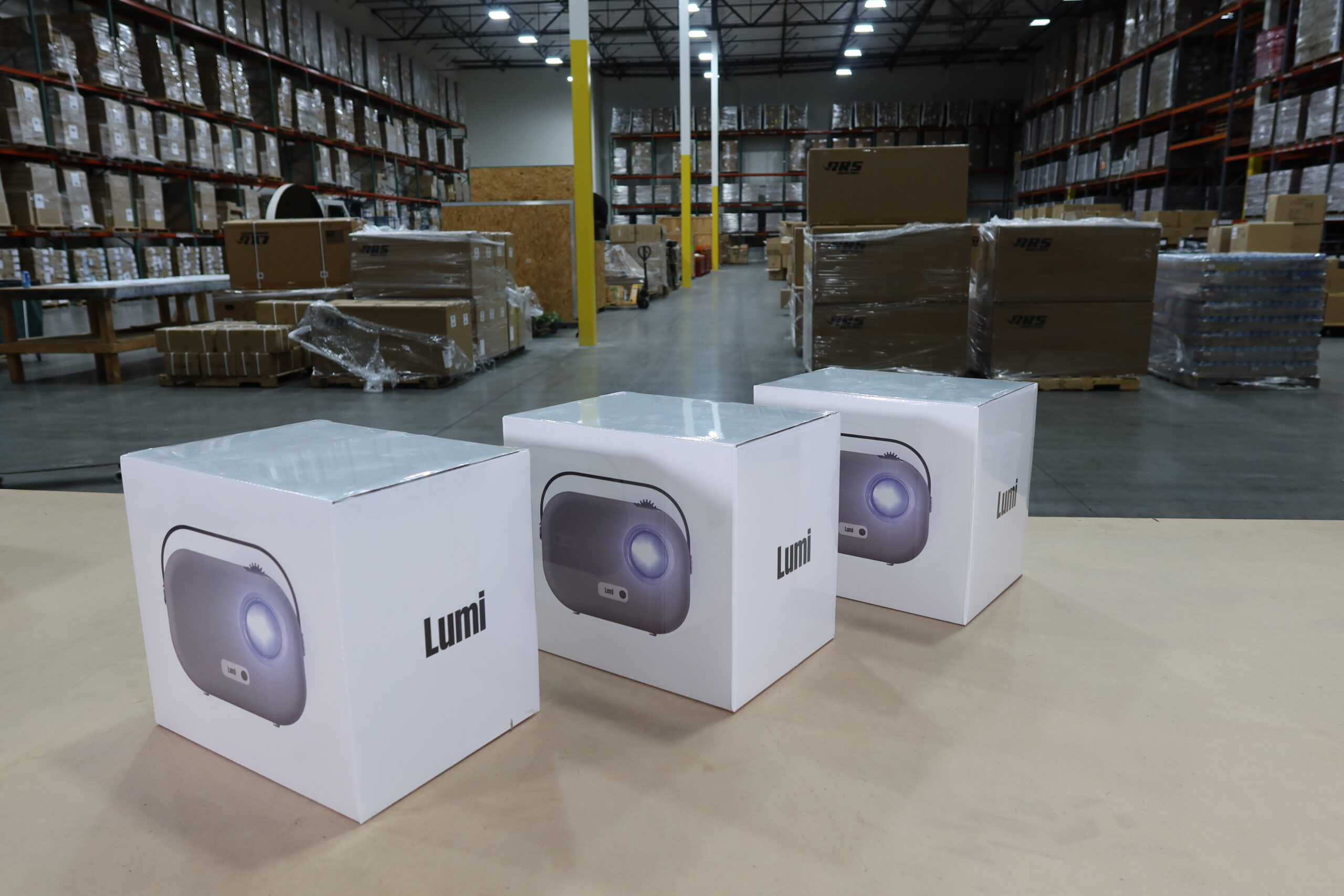 Three Lumi projectors in boxes on a warehouse table. Behind them is a warehouse made for 3PL work and tech brand logistics.